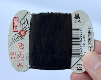 Op de afbeelding: Een klos zwart naaigaren gewikkeld om een bedrukte kartonnen kaart. De kaart bevat Japanse tekst, een vleugelafbeelding, een streepjescode en de tekst 40m. Het garen is 100% zijde, aangegeven op de kaart, en is gelabeld als nummer 9.
