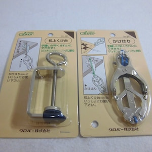 May include: Two packages of sewing tools. The package on the left contains a silver clamp with a blue base and a hook on top. The package on the right contains a silver clamp with a blue handle. Both packages have Japanese text on them.