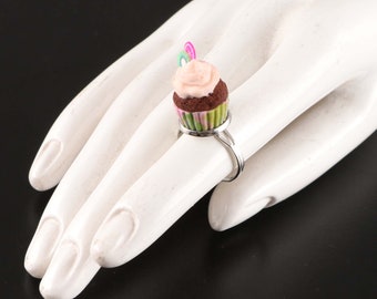 May include: A silver ring with a cupcake charm. The cupcake is brown with pink frosting and a green and pink swirl on top. The cupcake has a pink and green candy on top.