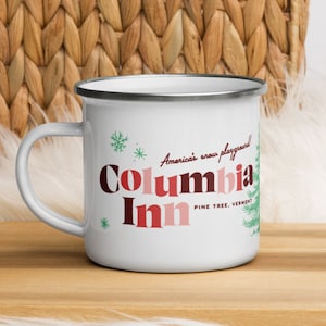 White Christmas inspired camper mug with a silver rim sitting on a wooden cutting board. The mug reads "Columbia Inn, Pine Tree, Vermont" with another line that says "America's snow playground".
