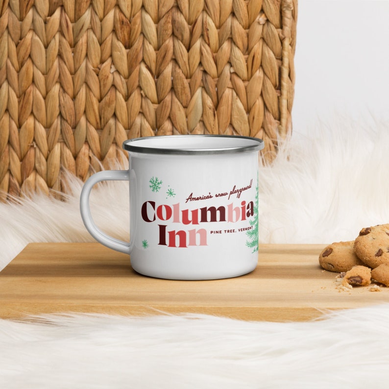 White Christmas inspired camper mug with a silver rim sitting on a wooden cutting board. The mug reads "Columbia Inn, Pine Tree, Vermont" with another line that says "America's snow playground".