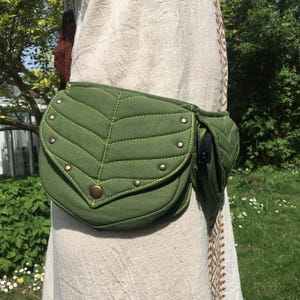 May include: A green leaf-shaped fanny pack with a small zippered pocket. The pack is made of a textured fabric with decorative stitching and metal studs. The bag is worn around the waist.