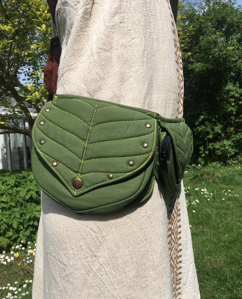 May include: A green leaf-shaped fanny pack with a small zippered pocket. The pack is made of a textured fabric with decorative stitching and metal studs. The bag is worn around the waist.
