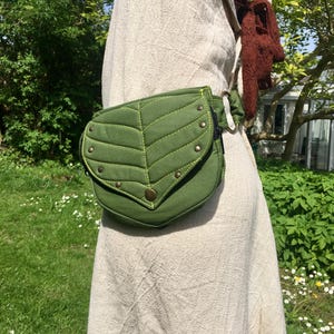 May include: A green leaf-shaped fanny pack with a flap closure and brass-colored rivets. The bag is made of a textured fabric and has a contrasting lime green stitching. The bag is worn around the waist.