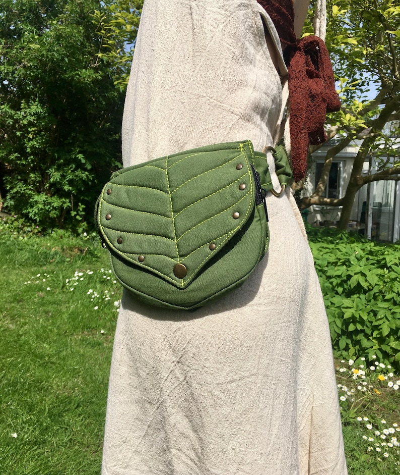 May include: A green leaf-shaped fanny pack with a flap closure and brass-colored rivets. The bag is made of a textured fabric and has a contrasting lime green stitching. The bag is worn around the waist.