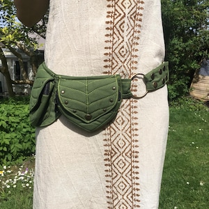 May include: A green quilted fanny pack with a leaf-shaped design and brass-colored rivets. The belt is olive green with brass studs and a large metal ring. The fanny pack is worn over a cream-colored dress with a brown geometric pattern.