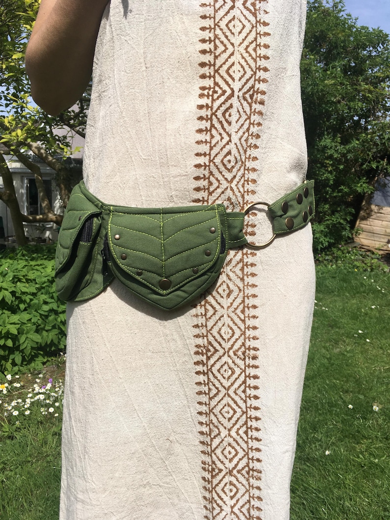 May include: A green quilted fanny pack with a leaf-shaped design and brass-colored rivets. The belt is olive green with brass studs and a large metal ring. The fanny pack is worn over a cream-colored dress with a brown geometric pattern.