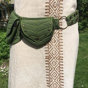 May include: A green quilted fanny pack with a leaf-shaped design on the front pocket. The belt features a brass ring and is secured with brass-colored studs. The fanny pack is worn around the waist over a cream-colored dress.