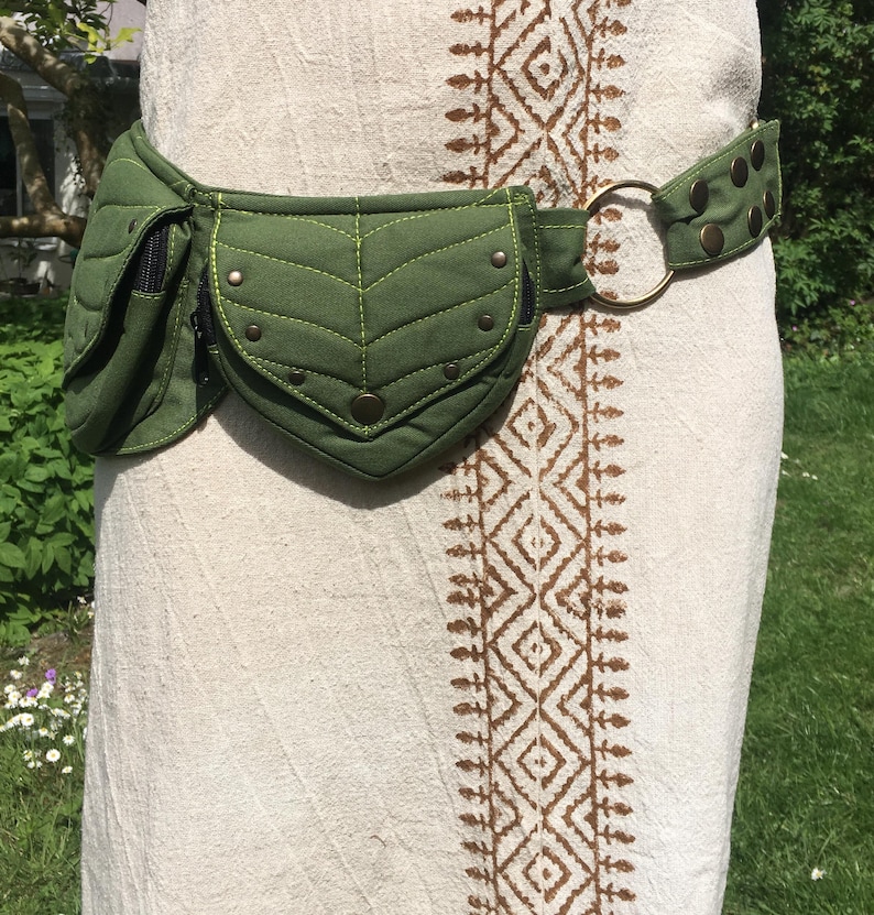 May include: A green quilted fanny pack with a leaf-shaped design on the front pocket. The belt features a brass ring and is secured with brass-colored studs. The fanny pack is worn around the waist over a cream-colored dress.