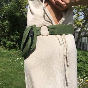 May include: A green quilted fanny pack with a black zipper and a matching green belt. The belt features a gold-colored circular buckle. The fanny pack is worn over a beige dress.