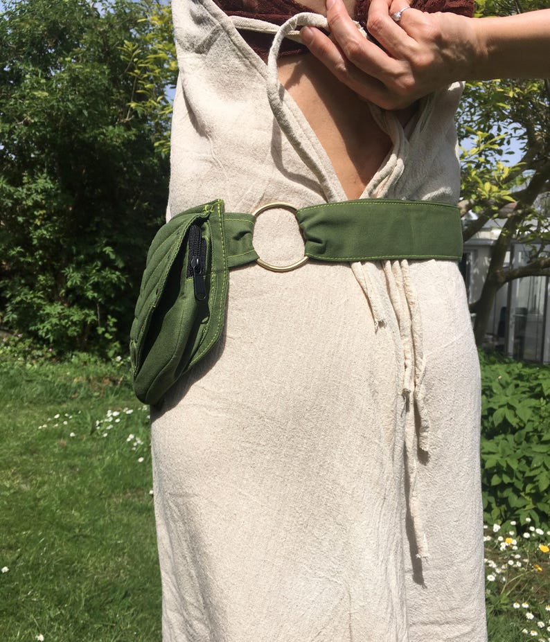 May include: A green quilted fanny pack with a black zipper and a matching green belt. The belt features a gold-colored circular buckle. The fanny pack is worn over a beige dress.