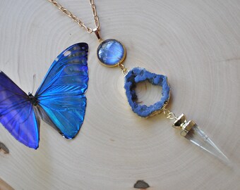 May include: A gold chain necklace with a blue agate pendant, a clear crystal point, and a round blue glass pendant. The necklace is displayed on a wooden surface with a blue morpho butterfly.