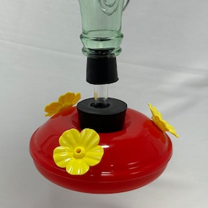 May include: A hummingbird feeder featuring a light green glass bottle neck attached to a bright red circular base. The base includes three yellow plastic flower-shaped feeding ports with small central openings and a black rubber stopper.