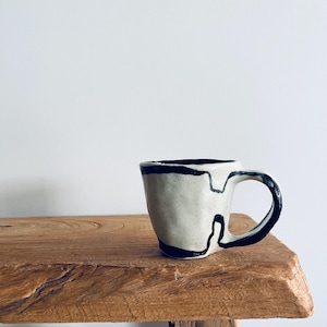 May include: A cream-coloured ceramic mug with a black abstract design. The mug has a rounded body, a curved handle, and a black rim. It sits on a rustic wooden surface, highlighting its unique, artistic design. The mug is likely handmade.