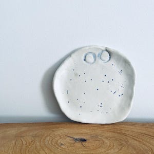 May include: A handmade, off-white ceramic plate with a whimsical design. The plate features two raised, blue circles near the top and scattered blue speckles across the surface. The plate is irregularly shaped and sits on a wooden surface.