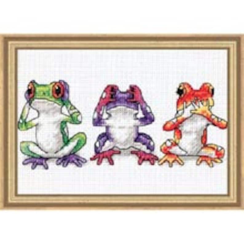 May include: Three frogs, each with a different color scheme, are depicted in a cross-stitch pattern. The frog on the left is green with purple spots, the middle frog is purple with red spots, and the frog on the right is red with orange spots. Each frog is covering its eyes, ears, and mouth with its hands, representing the "See no evil, hear no evil, speak no evil" proverb.