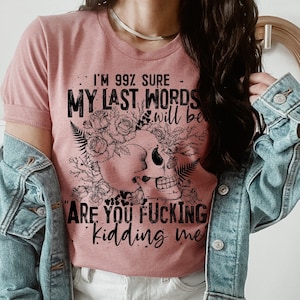 May include: Dusty rose t-shirt with a skull graphic surrounded by flowers and ferns. The text reads "I'm 99% sure my last words will be 'Are you fucking kidding me'". The shirt is paired with a denim jacket and white shorts.