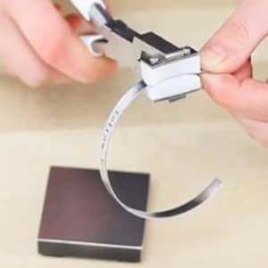May include: A person using a metal bending tool with white handles to shape a thin, silver metal strip. The strip is curved, and a small, square metal block sits below. The tool is silver and white.