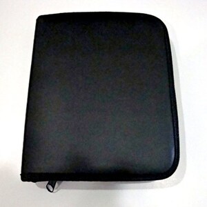 May include: A black zippered case, likely for documents or a tablet, is shown against a white background. The case is rectangular with rounded corners and a black zipper closure. The material appears to be a textured, matte finish.