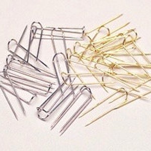 May include: A collection of silver and gold-colored pins, likely for sewing or crafting, are scattered on a white surface. The pins have a distinctive U-shaped design with a pointed end.