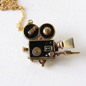 Op de afbeelding: Een gouden ketting met een vintage-stijl camera hanger. De camera is gemaakt van messing en heeft zwarte details. De camera heeft twee ronde lenzen en een klein handvat aan de zijkant.
