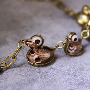 May include: A gold chain bracelet with three small duck charms. Each duck charm is made of a brown and white material and has a skeletal design. The ducks have large, black eyes.