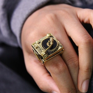 May include: A gold-colored ring shaped like a vintage turntable. The ring features a detailed design of a record player with a black record and a tone arm. The ring is worn on a finger.