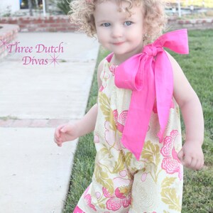 May include: A young child wearing a floral print romper with pink accents and a large pink bow. The romper features a cream background with pink and gold floral patterns, pink polka dot ruffles, and a matching bow.