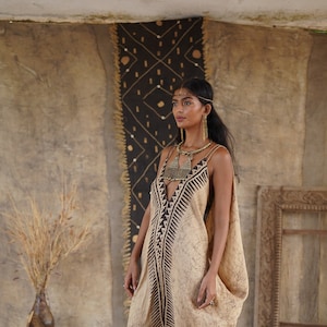 May include: A beige maxi dress with a V-neckline and a black geometric pattern. The dress has thin straps and a flowing silhouette. The model is wearing gold jewelry, including a necklace and earrings. The background features a patterned wall and a decorative rug.