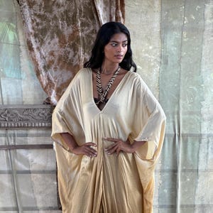 May include: A long, flowing, ombre kaftan with a V-neckline. The kaftan is made of a silky fabric and is a light beige color that fades to a brown color at the bottom.