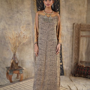 May include: A long, flowing dress with a brown and beige abstract pattern. The dress has a scoop neck and cut-out shoulders. The model is wearing a silver necklace with a tassel.