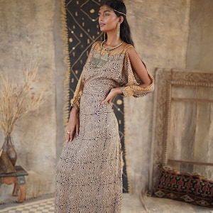 May include: A woman wearing a long, brown and gold patterned dress with cut-out shoulders. She is standing in front of a wall with a black and white patterned rug hanging on it.