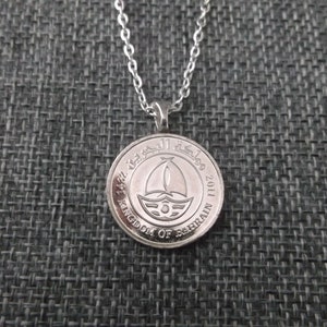 May include: Silver necklace with a round pendant featuring the coat of arms of the Kingdom of Bahrain and the year 2011.