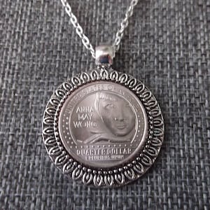 May include: Silver necklace with a round pendant featuring a silver coin with the image of Anna May Wong and the text "United States of America" and "Quarter Dollar" on it.
