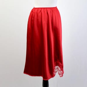 May include: A red satin slip skirt displayed on a mannequin. The skirt has a mid-length hemline and a delicate lace trim at the bottom. The mannequin is on a black stand against a white background.