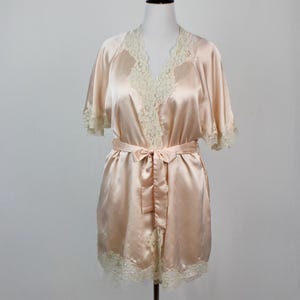 May include: A blush-colored satin robe with ivory lace trim. The short-sleeved robe features a matching tie belt and lace detailing along the neckline and hem. The robe is displayed on a black mannequin stand against a white background.