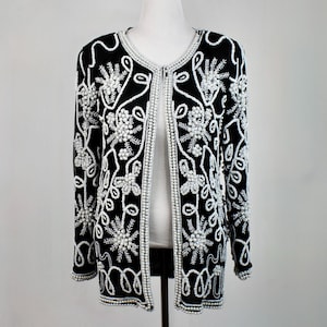 May include: Black cardigan with white floral and swirl beadwork. The open front jacket features a round neckline and long sleeves. The white beading creates a striking contrast against the black fabric, creating a vintage look.