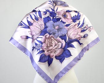 May include: Square silk scarf with a floral design. The scarf features a white background with a large bouquet of pink, purple, and blue flowers. The edges are trimmed with a lavender border. The scarf is displayed on a mannequin.