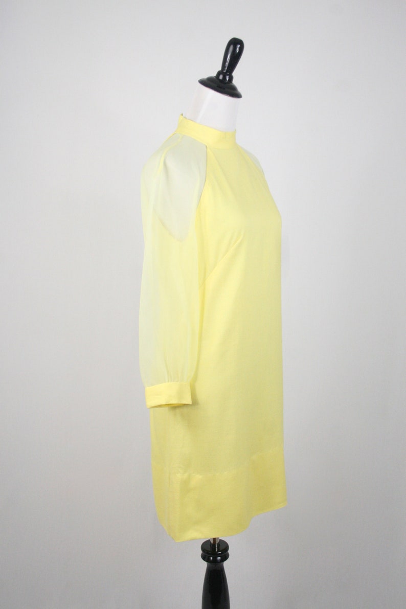 May include: A yellow dress with sheer sleeves. The dress has a high neckline and a simple, straight silhouette.