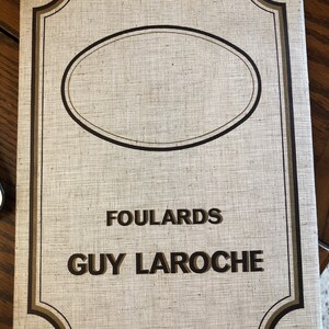 May include: A white book cover with a brown border and a brown oval in the center. The text "FOULARDS GUY LAROCHE" is printed in brown letters.