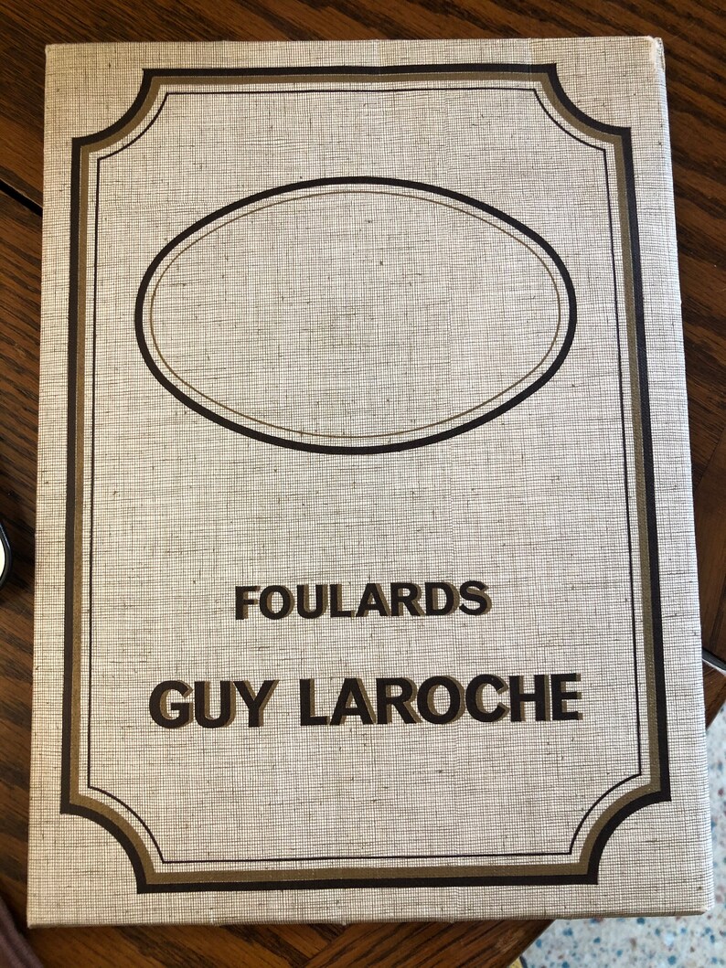 May include: A white book cover with a brown border and a brown oval in the center. The text "FOULARDS GUY LAROCHE" is printed in brown letters.
