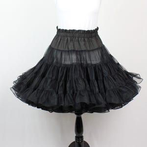 May include: A black petticoat with multiple layers of ruffled fabric, displayed on a black mannequin stand. The petticoat has an elastic waistband and a full, flared silhouette. The garment is designed to add volume to skirts.