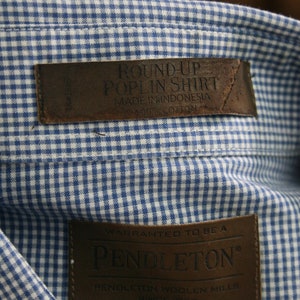 May include: A blue and white gingham shirt with a brown leather label that reads "Round-Up Poplin Shirt Made in Indonesia 100% Cotton" and a brown leather label that reads "Warranted to be a Pendleton Pendleton Woolen Mills Since 1883" with a size "S" tag.