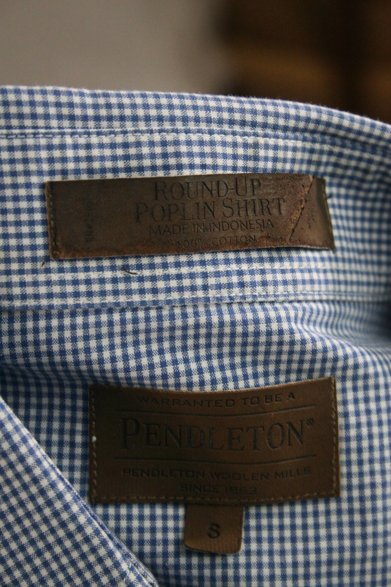 May include: A blue and white gingham shirt with a brown leather label that reads "Round-Up Poplin Shirt Made in Indonesia 100% Cotton" and a brown leather label that reads "Warranted to be a Pendleton Pendleton Woolen Mills Since 1883" with a size "S" tag.