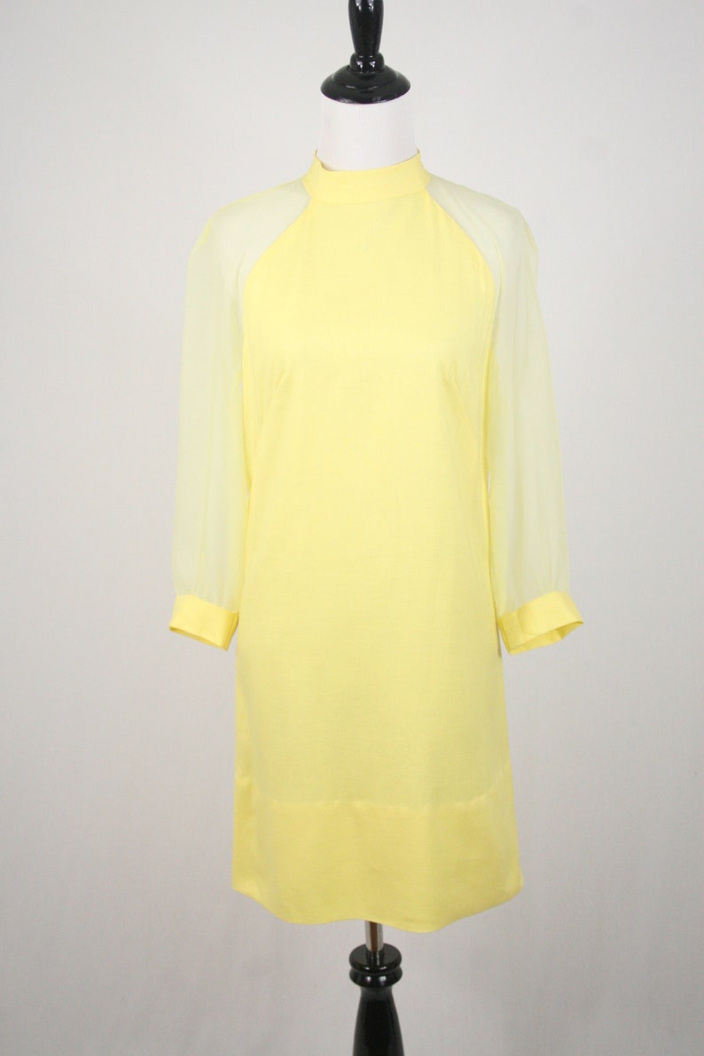 May include: A yellow dress with sheer sleeves. The dress has a high neck and a straight silhouette.