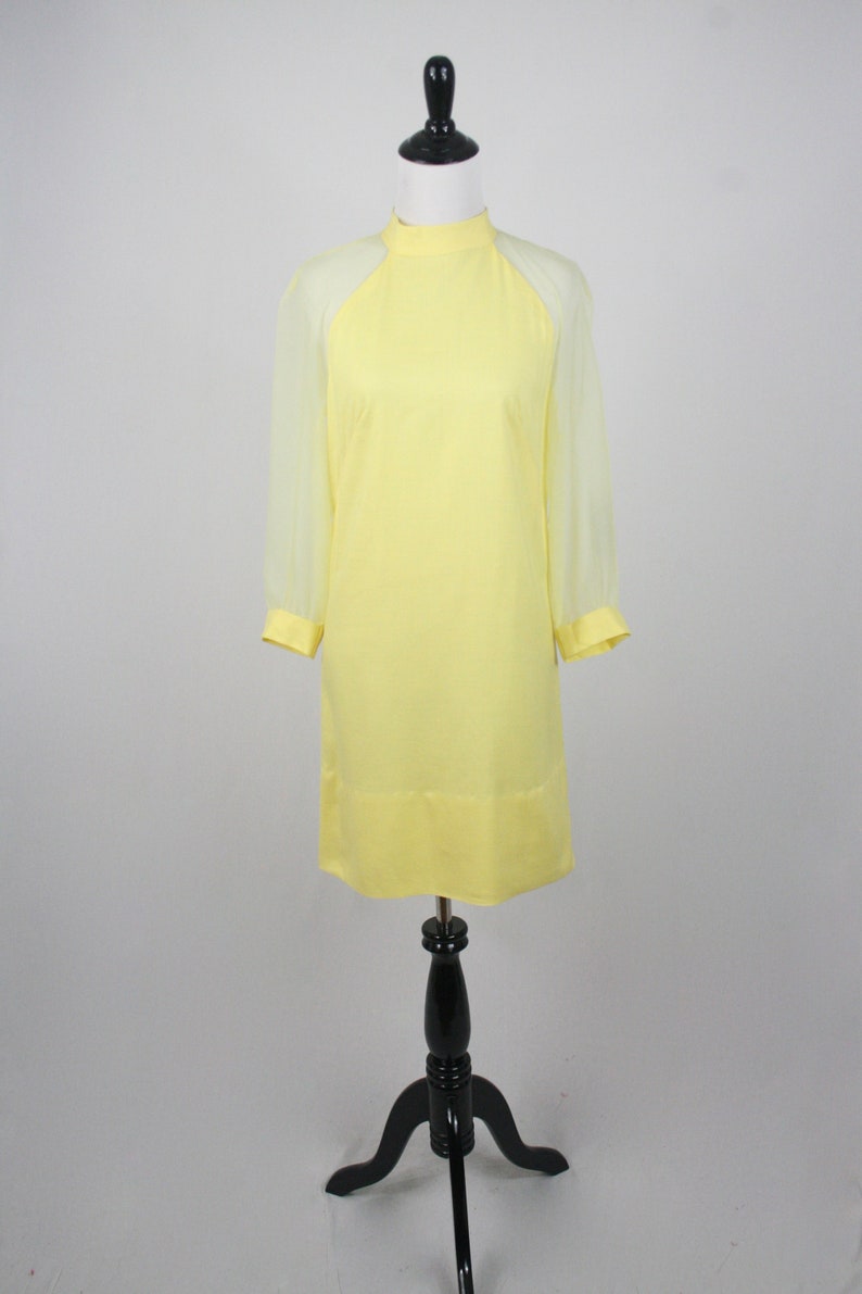 May include: A yellow dress with sheer sleeves. The dress has a high neck and a simple, straight silhouette.