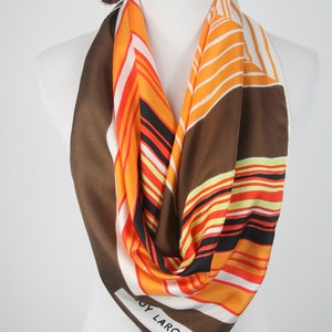 May include: A white silk scarf with brown, orange, and black stripes. The scarf has the text "Guy Laroche Paris" printed on it.