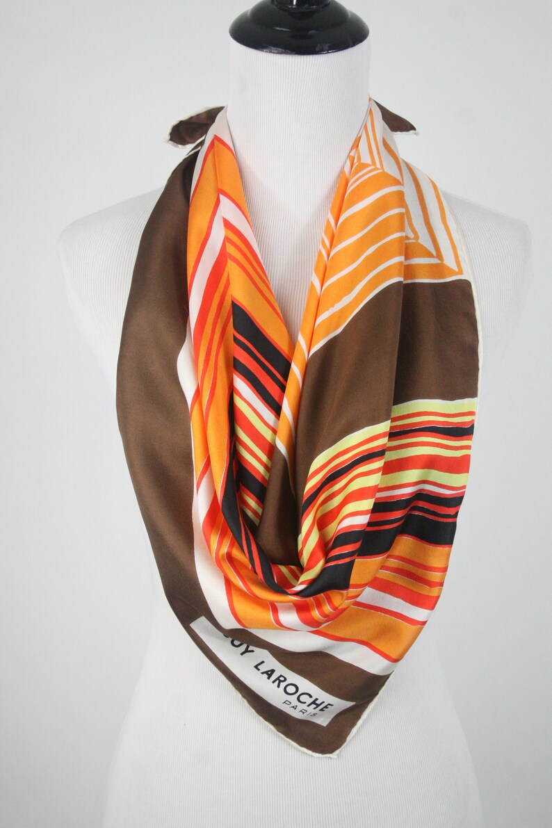 May include: A white silk scarf with brown, orange, and black stripes. The scarf has the text "Guy Laroche Paris" printed on it.