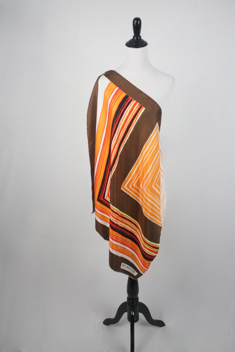 May include: A brown, orange, and white striped scarf with a geometric pattern. The scarf is draped over a white mannequin and has a "Guy Laroche" label.