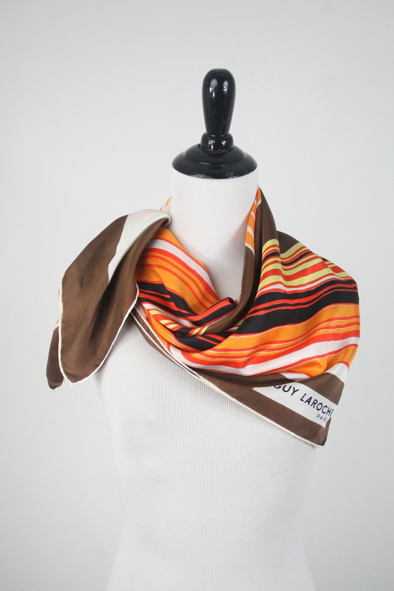 May include: A brown, orange, white, and black striped silk scarf with the words 'GUY LAROCHE PARIS' printed on the bottom right corner.